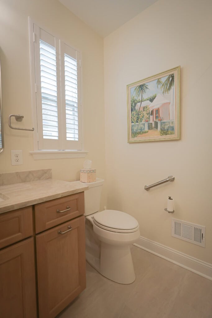 a clean, modern bathroom with light wood cabinets, a marble countertop, a white toilet, a wall mounted toilet paper holder, a vent, a window with shutters, and a framed painting of a tropical house on the wall.
