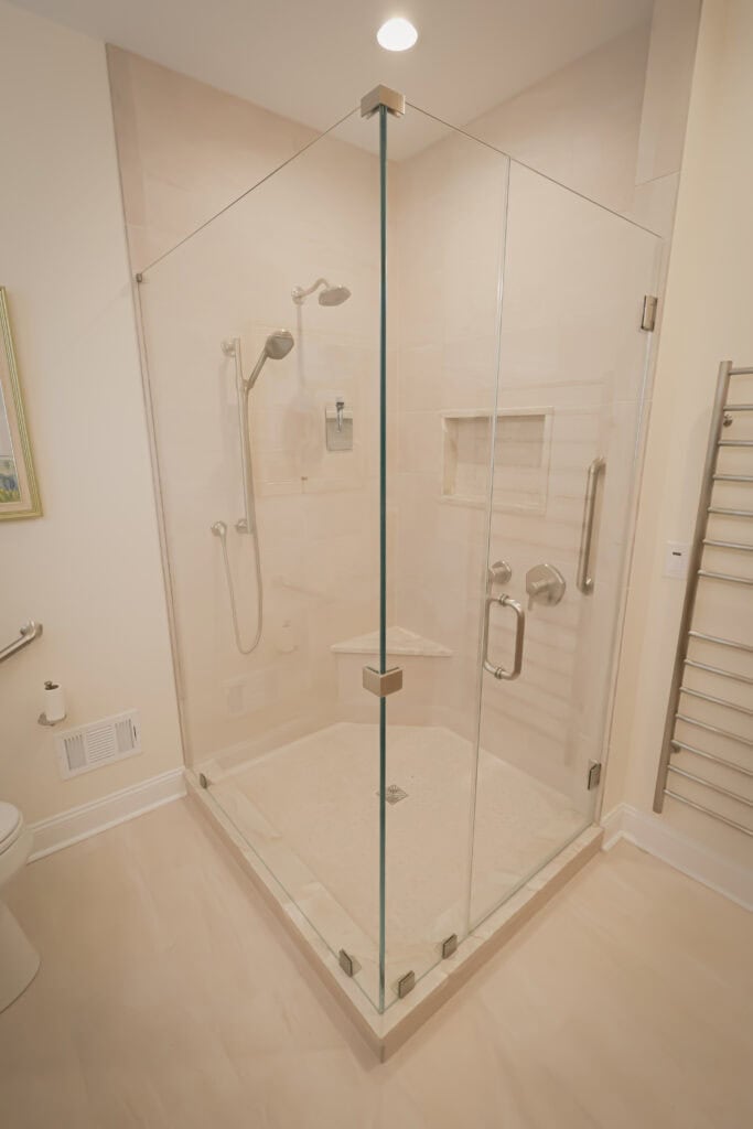 a modern, corner glass shower with chrome fixtures, a built in shelf, and a small bench. the shower is in a beige tiled bathroom with a towel warmer and toilet visible nearby.