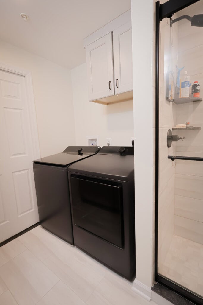 a modern laundry room with a black washer and dryer set, white wall cabinets above, light tile flooring, and a glass shower enclosure to the right.