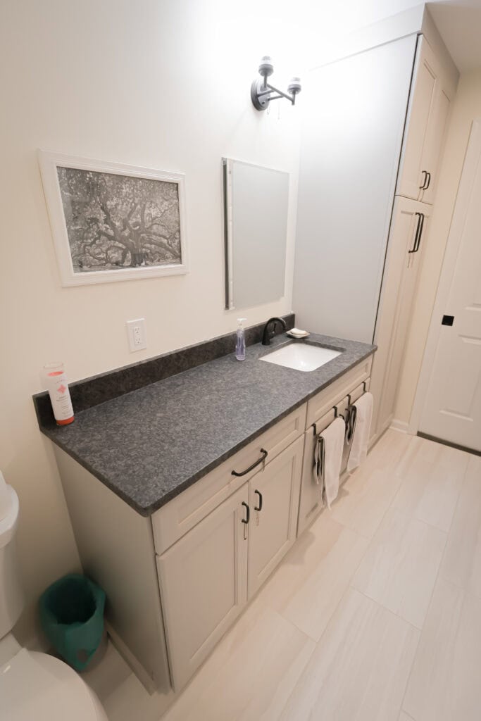 a modern bathroom with light colored cabinets, a dark countertop, a sink, a mirror, wall art, white towels hanging from cabinet handles, and a tall storage cabinet beside the vanity.