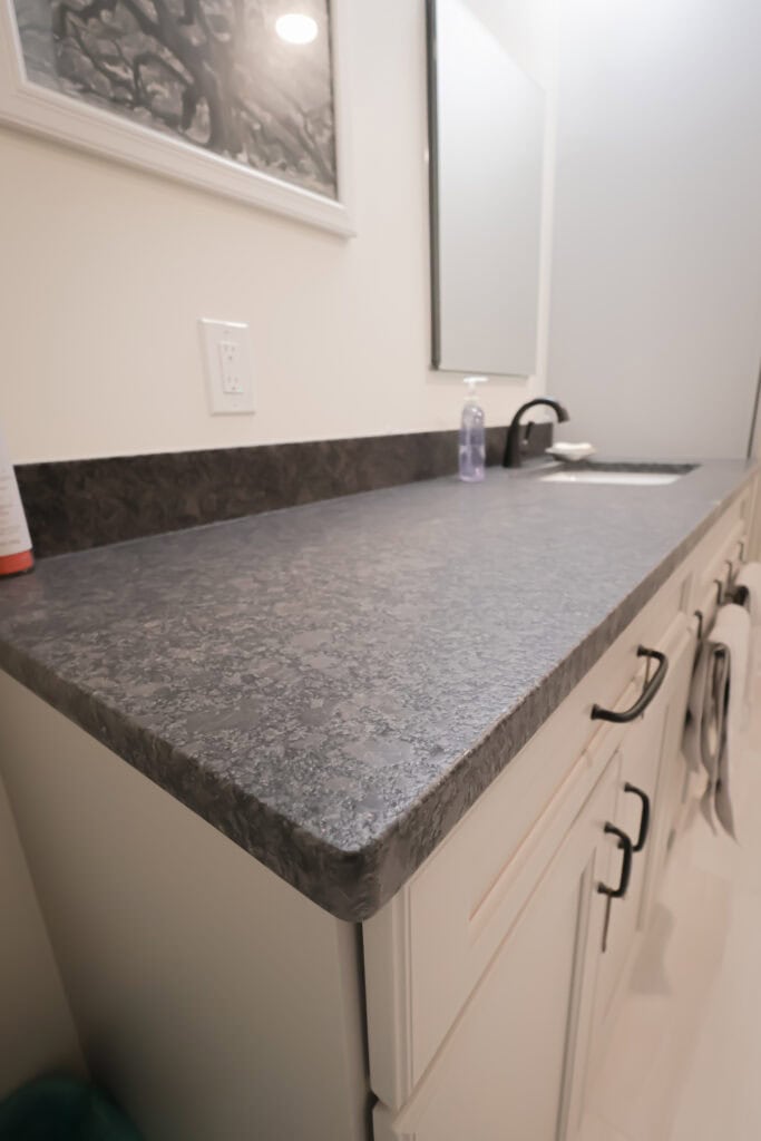 a bathroom countertop with a dark, textured surface, white cabinets with black handles, a black faucet, a soap dispenser, and a towel hanging on the cabinet. a mirror and wall art are visible in the background.