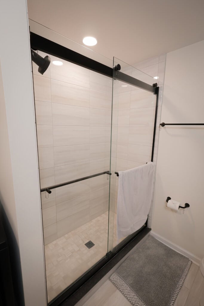 modern walk in shower with sliding glass doors, black hardware, light beige tile walls, a towel hanging on the door, a gray bath mat on the floor, and a toilet paper holder mounted to the white wall.