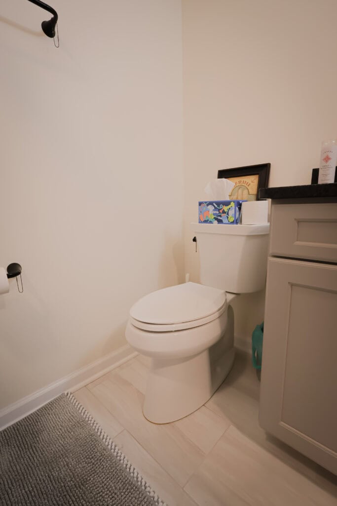 a bathroom with a white toilet next to a beige cabinet, a box of tissues and framed item on top of the toilet tank, toilet paper on a holder, and a gray bath mat on the light tile floor.