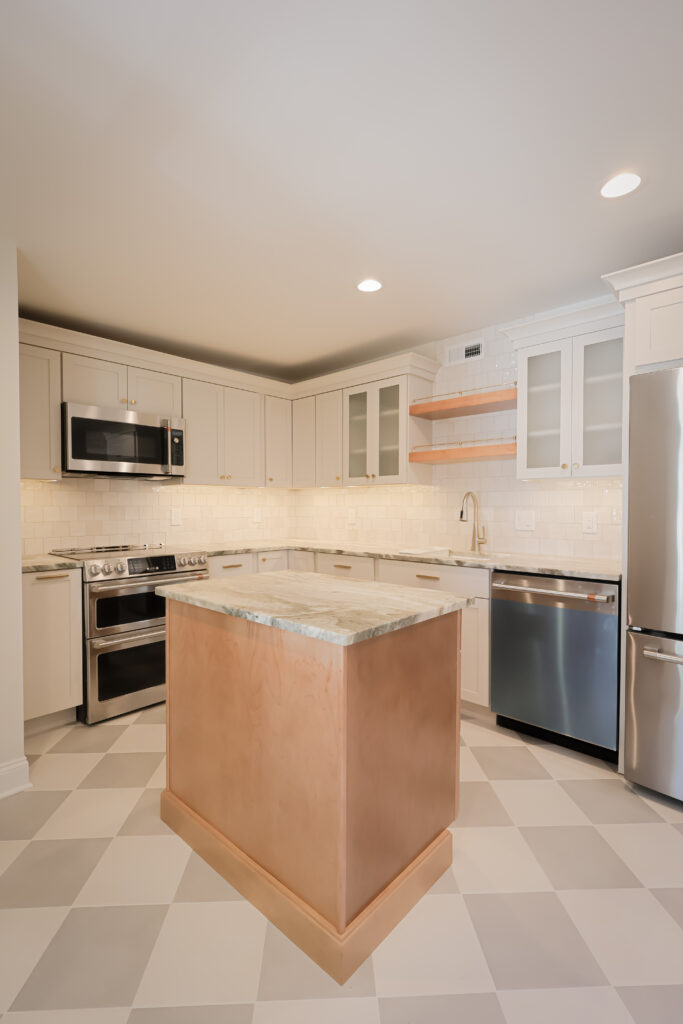 modern kitchen with light cabinets, stainless steel appliances, a marble topped island with a copper base, open wooden shelves, tile backsplash, and checkered light tile flooring.
