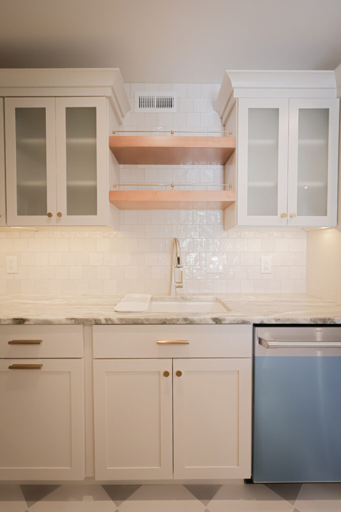 modern kitchen with white cabinets, marble countertop, a sink with a high arc faucet, wooden open shelves, glass cabinet doors, and a blue dishwasher, all set against a white tile backsplash.