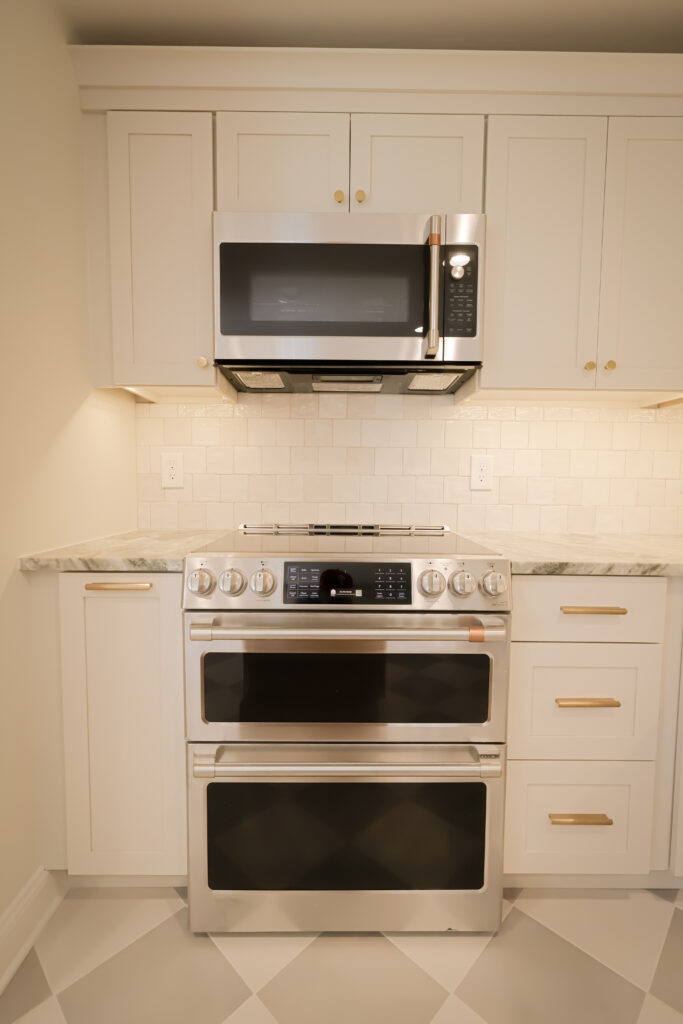 modern kitchen with white cabinets, gold handles, a built in stainless steel microwave, and a double oven. the backsplash is white tile, and the countertop is light colored marble. the floor has a diamond pattern.