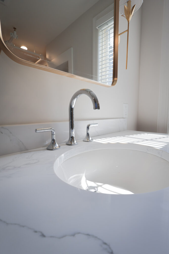 close up of a modern bathroom sink with a marble countertop, chrome faucet, and handles. sunlight filters in through window blinds, casting shadows on the counter.