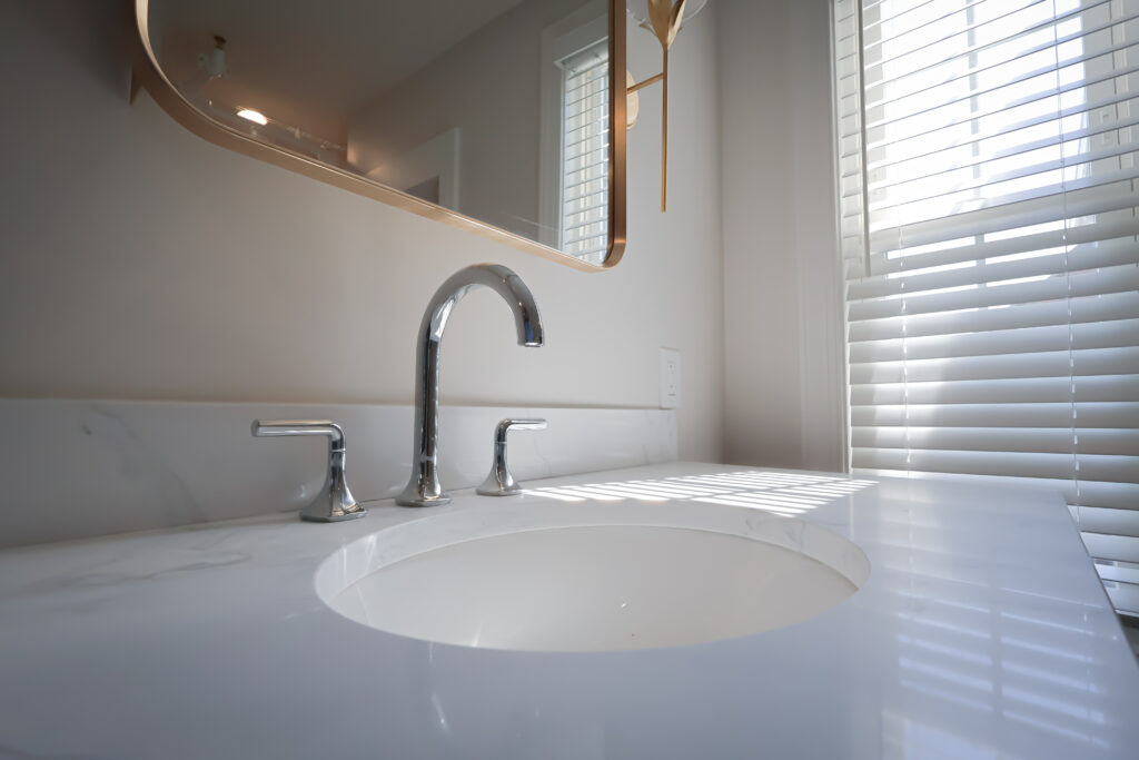 close up of a modern bathroom sink with a chrome faucet and handles, a white countertop, a large mirror, and sunlight streaming through white blinds on the window.