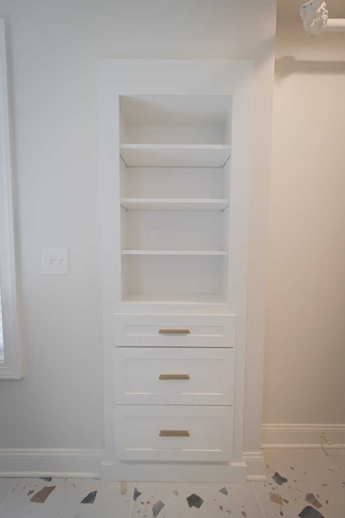 built in white shelving unit with three open shelves above and three drawers below, each with a gold handle. the unit is set into a light colored wall next to a window and above a speckled floor.