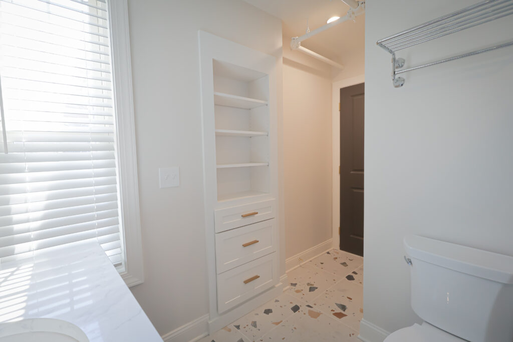 bright bathroom with white shelving, white toilet, terrazzo tile floor, and a counter with a sink. there are blinds on the window and a dark colored door in the back.