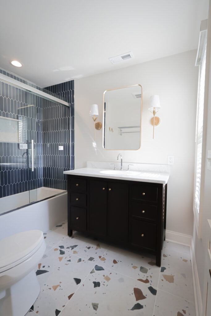 modern bathroom with dark vanity, gold hardware, a rectangular mirror, white sink, terrazzo floor, and a bathtub with glass door and blue geometric tiles on the wall.