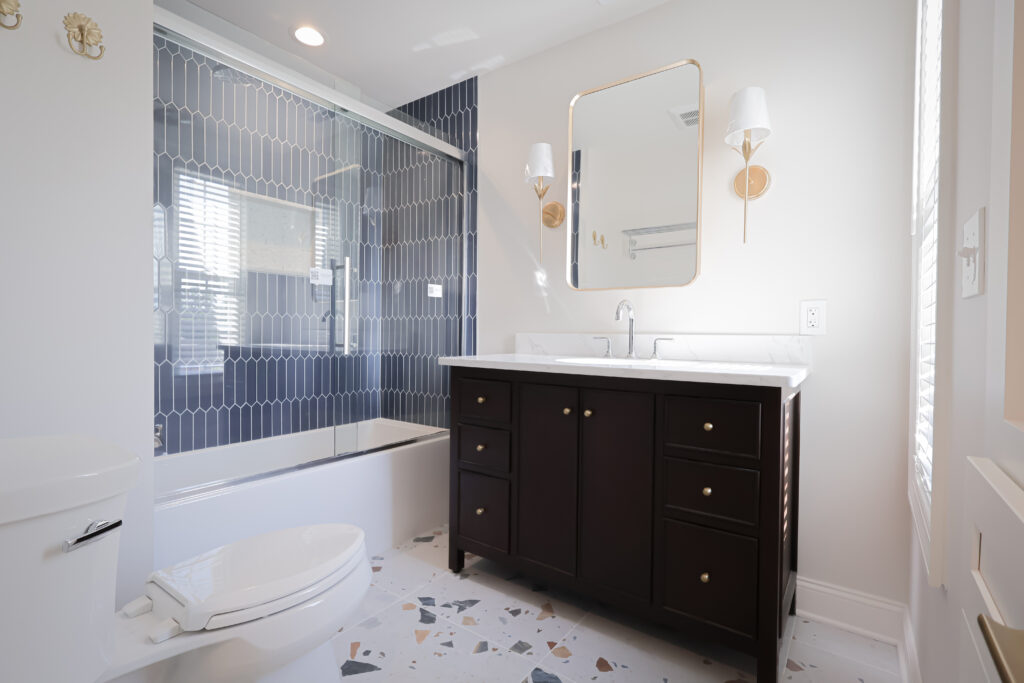 modern bathroom with a dark vanity, white sink, large mirror, wall sconces, toilet, and glass enclosed tub with blue geometric tile. terrazzo style flooring and natural light from a window.