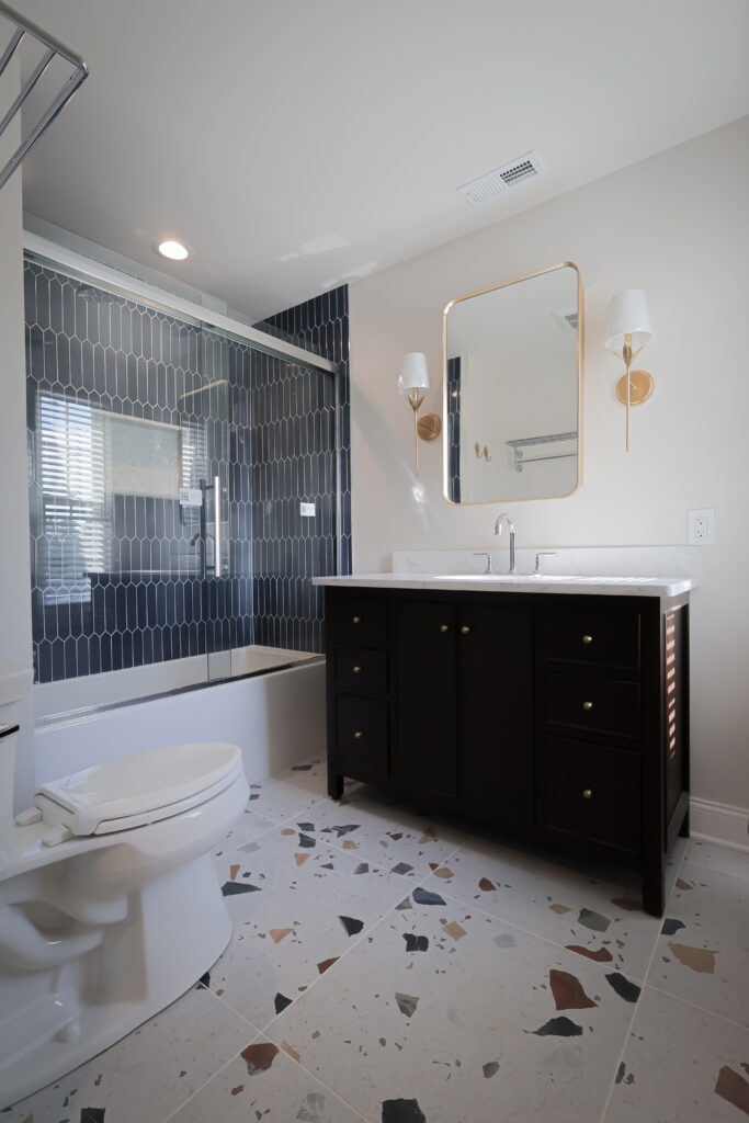 modern bathroom with a terrazzo floor, dark vanity with white countertop, oval mirror, wall sconces, and a bathtub with blue geometric tile and glass shower door. light walls and natural light from window.