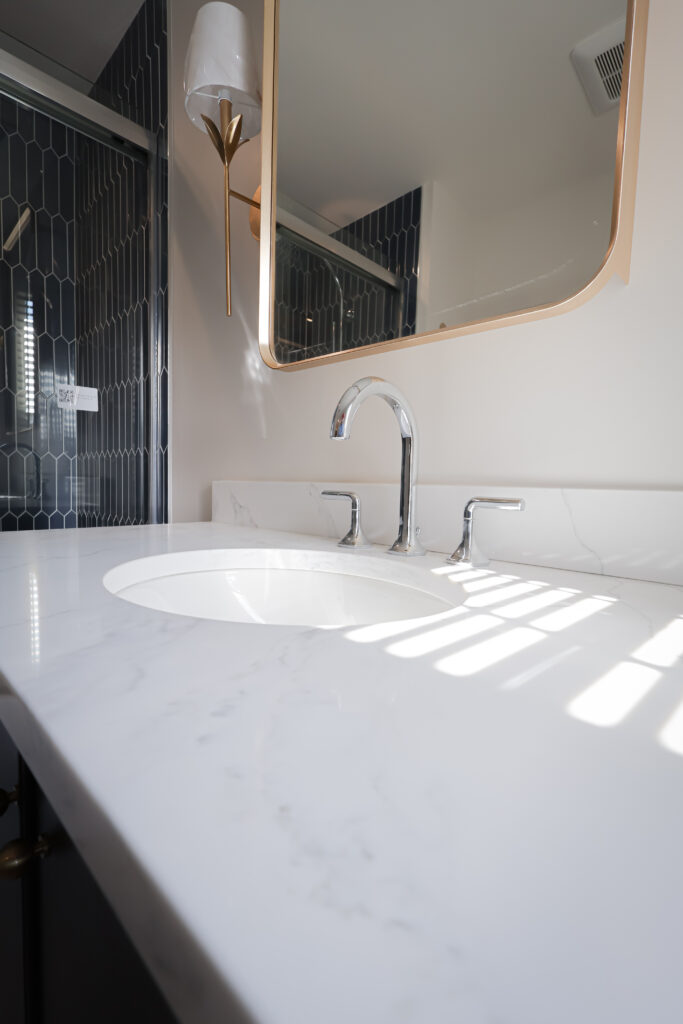 modern bathroom with a white marble countertop, oval sink, chrome faucet, gold framed mirror, and geometric dark blue tile wall in the background. sunlight creates striped shadows on the counter.