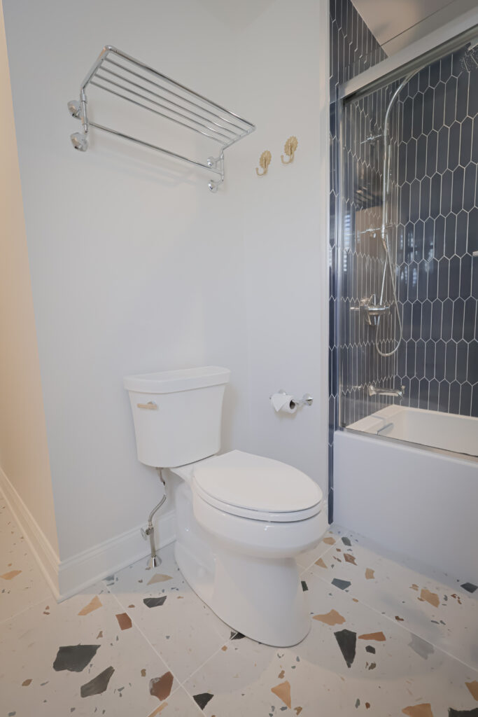 modern bathroom with a white toilet, chrome towel rack and hooks, toilet paper holder, terrazzo flooring, and a bathtub with dark blue hexagonal tile and a glass shower door.