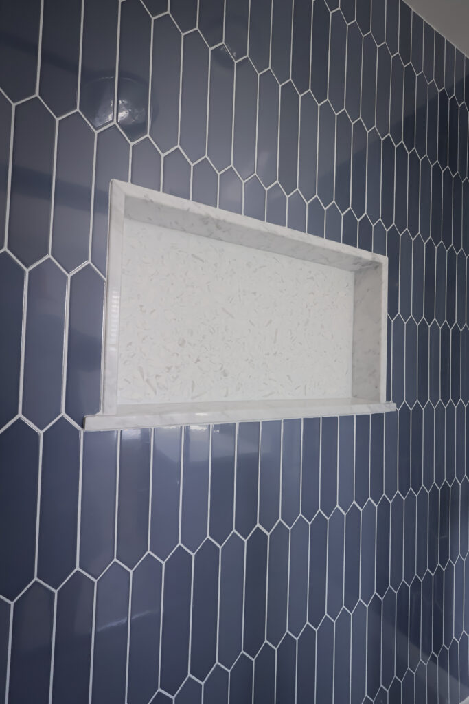 a white rectangular shower niche with a marble frame is set into a wall covered with glossy, dark blue hexagonal tiles arranged vertically.