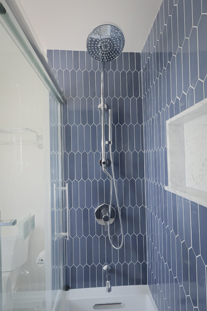 modern shower with tall blue hexagonal tiles, a chrome showerhead, and a matching handheld wand. there is a recessed shelf in the tiled wall and part of a glass shower door is visible on the left side.