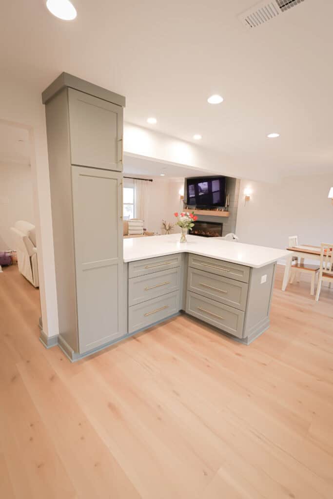 Modern kitchen corner with light green cabinets, brass handles, white countertops, and wood flooring. A vase with flowers sits on the counter. The background shows a living area with a TV, fireplace, and dining table.