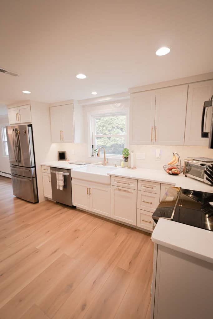 Bright modern kitchen with white cabinets, stainless steel appliances, farmhouse sink, wood floors, and a window above the sink letting in natural light. Countertops are uncluttered, with a few items and a fruit basket visible.