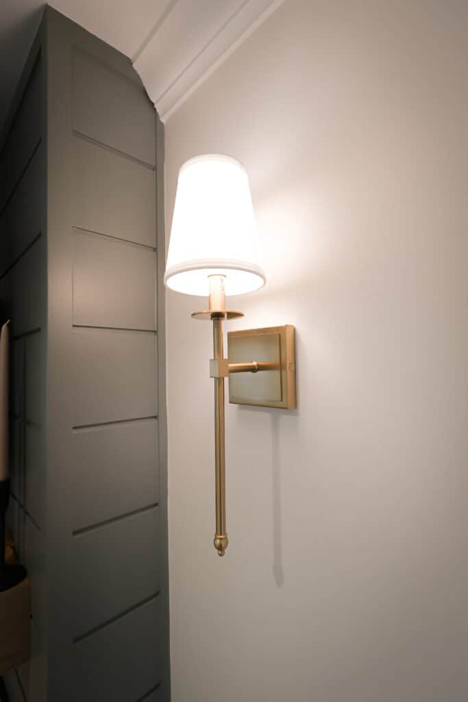 A modern brass wall sconce with a white lampshade is mounted on a white wall next to a gray paneled section. The light is turned on, emitting a soft, warm glow.