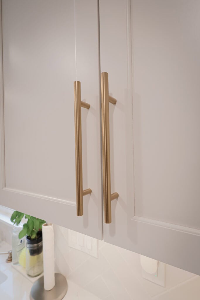 Close-up of two beige kitchen cabinet doors with long, gold vertical handles. In the background, a white countertop with a potted plant, a paper towel holder, and electrical outlets is visible.