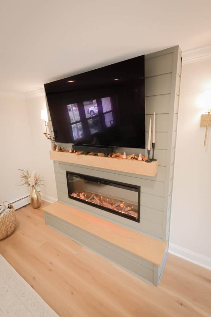 A modern living room features a wall-mounted flat-screen TV above a sleek electric fireplace with a wooden mantel decorated with small ornaments and candles. Light wood flooring and soft lighting enhance the cozy ambiance.