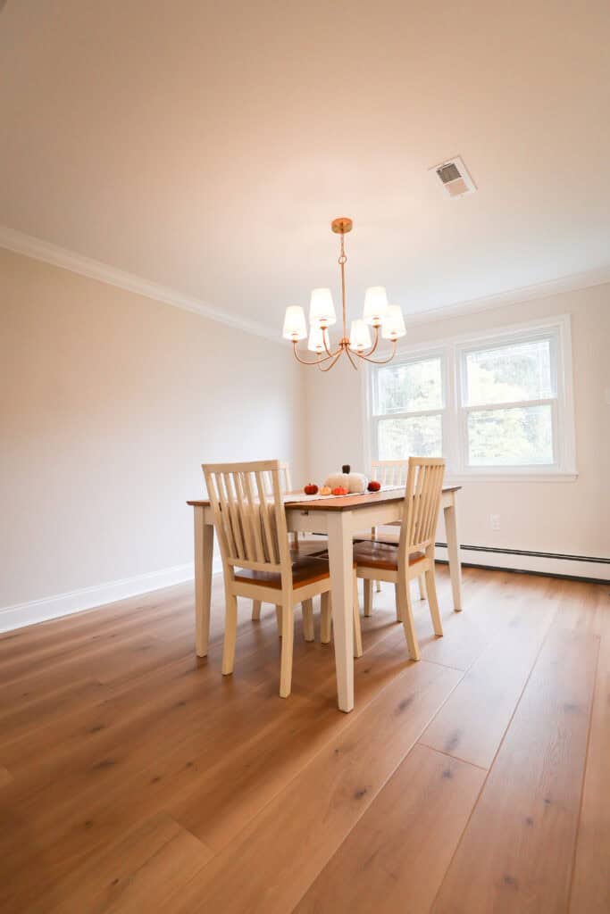 A simple dining room with a wooden table and four chairs, a modern chandelier above, light wood flooring, and two windows letting in natural light. A small centerpiece decorates the table.