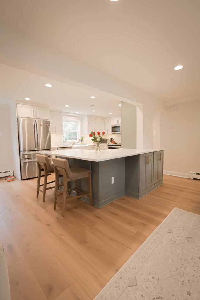 A modern kitchen with light wood floors, a large island with a white countertop and gray base, three cushioned barstools, stainless steel appliances, white cabinets, and a vase of flowers on the island.