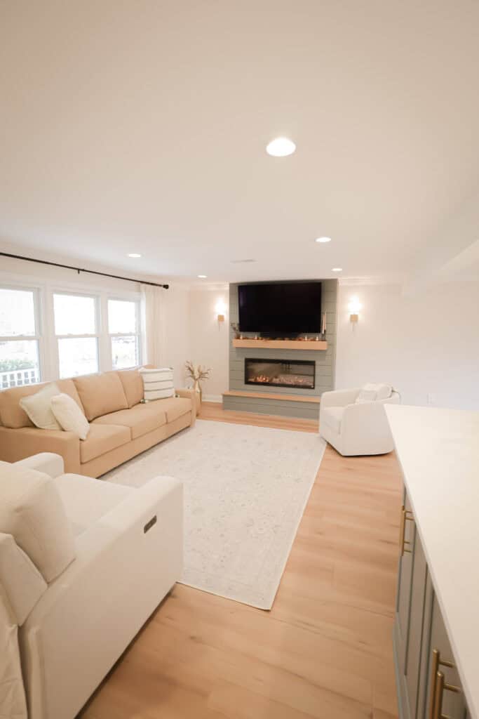 A bright, modern living room with light wood floors, beige couch, white armchair, large rug, wall-mounted TV above a fireplace, and large windows allowing natural light.
