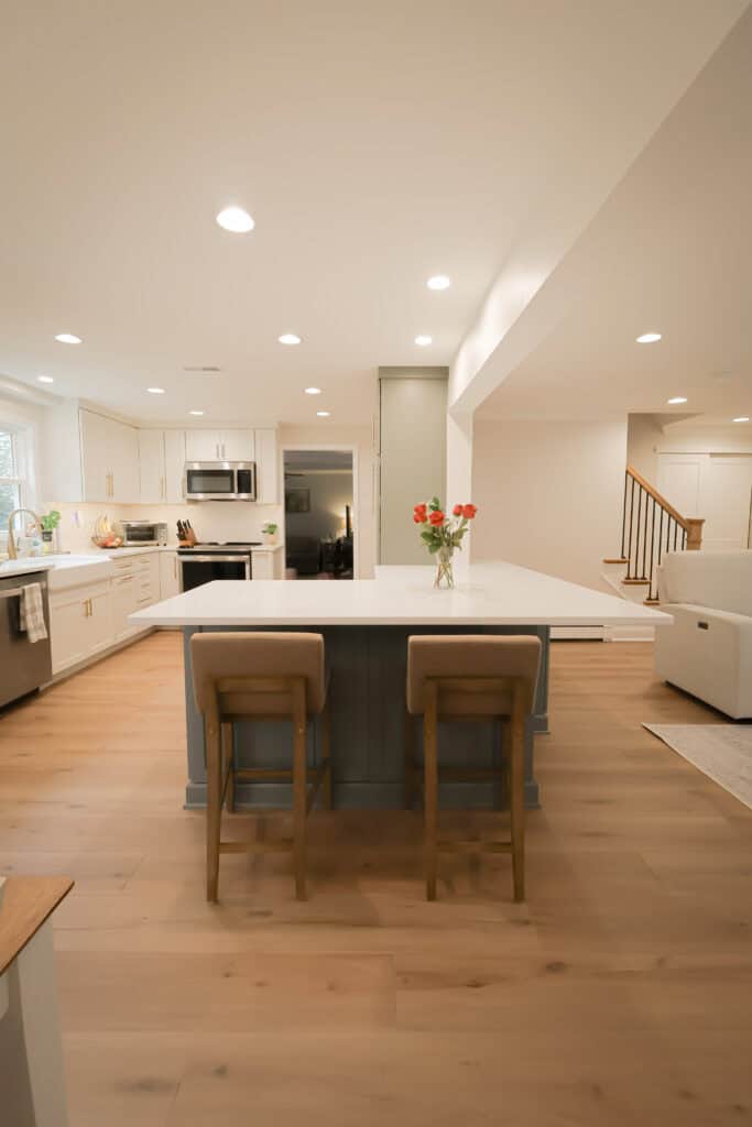 A modern, spacious kitchen with an island featuring two cushioned stools, white cabinets, stainless steel appliances, and hardwood floors. Fresh flowers in a vase sit on the island, and stairs are visible in the background.
