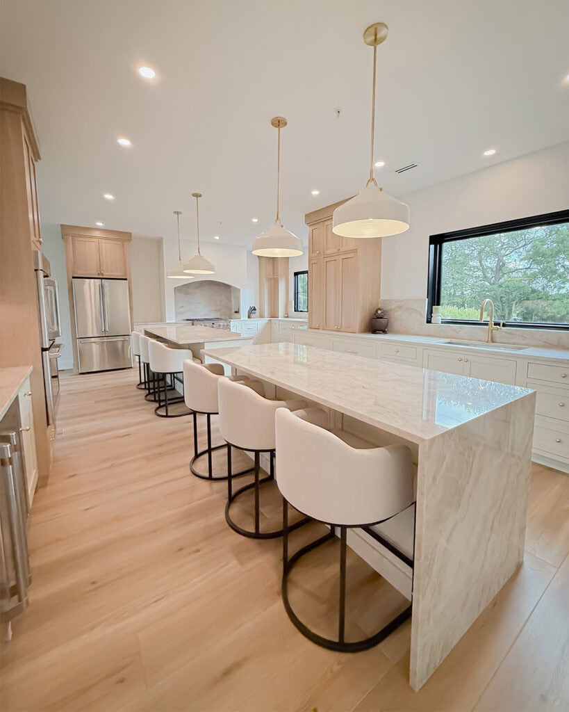 Modern kitchen with light wood floors, white marble island with four cushioned barstools, gold pendant lights, built-in appliances, and large window overlooking greenery.
