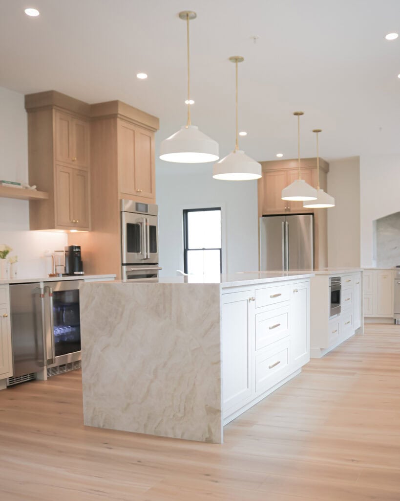 A modern kitchen with a large marble island, white cabinets, light wood accents, stainless steel appliances, pendant lights, and light wood flooring. The space is bright and open with minimalist decor.
