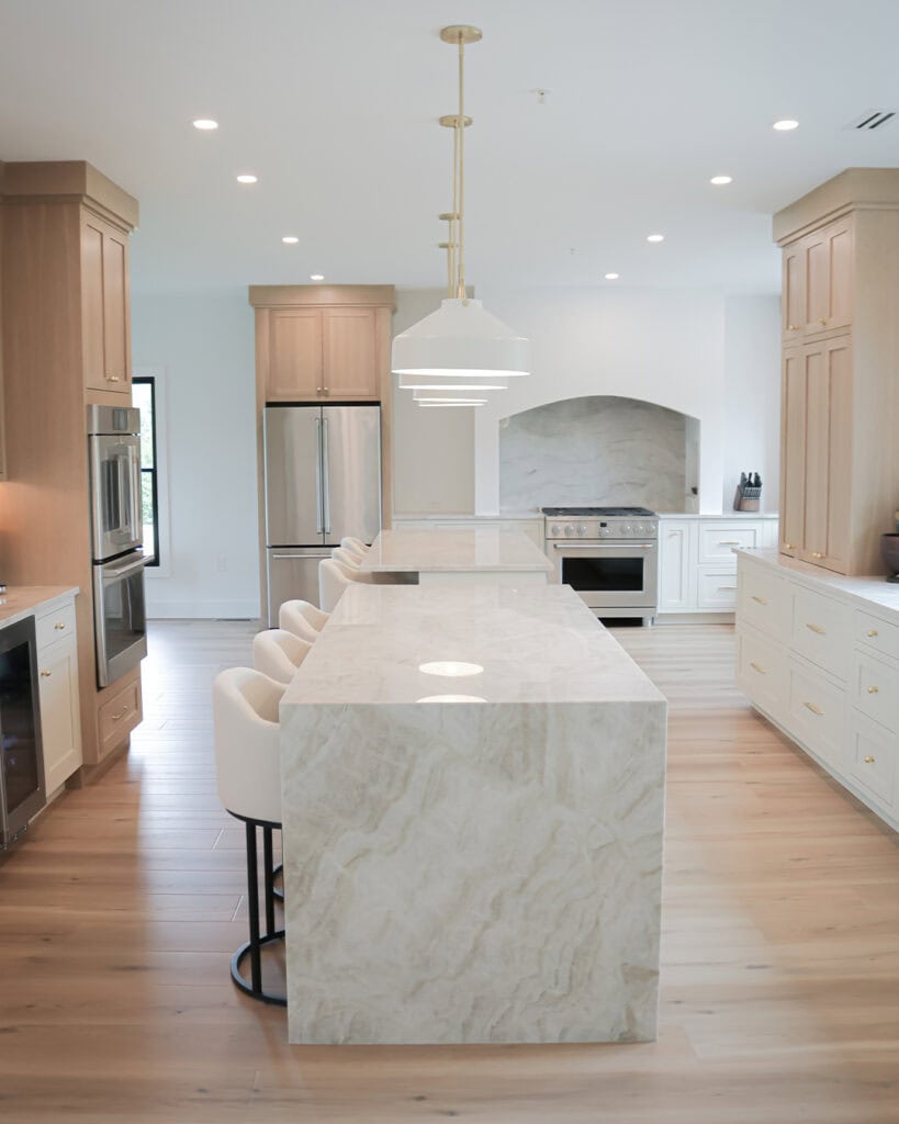 A modern kitchen with light wood cabinets, marble countertops and island, stainless steel appliances, white cushioned bar stools, pendant lights, and light wood flooring.