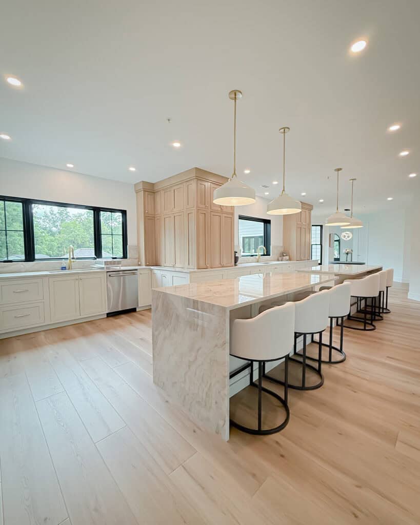 Spacious modern kitchen with light wood floors, large marble island with four white barstools, pendant lights, and light wood cabinetry. Black-framed windows provide ample natural light.