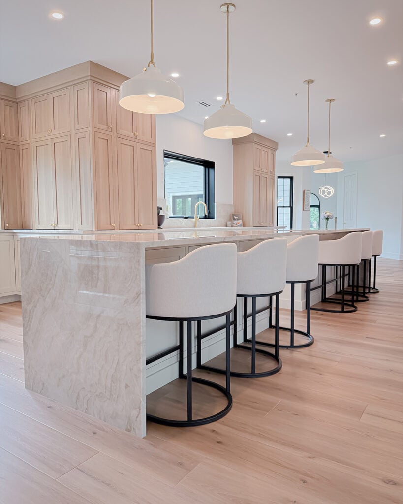 A modern kitchen with light wood cabinets, a large marble island, four white upholstered bar stools with black bases, pendant lights overhead, and light wood flooring. Natural light fills the space.