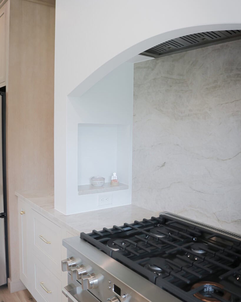 A modern kitchen features a stainless steel gas stove beneath a white arch with a marble backsplash. A small recessed shelf holds a bowl and a bottle, and there is an outlet below the shelf.