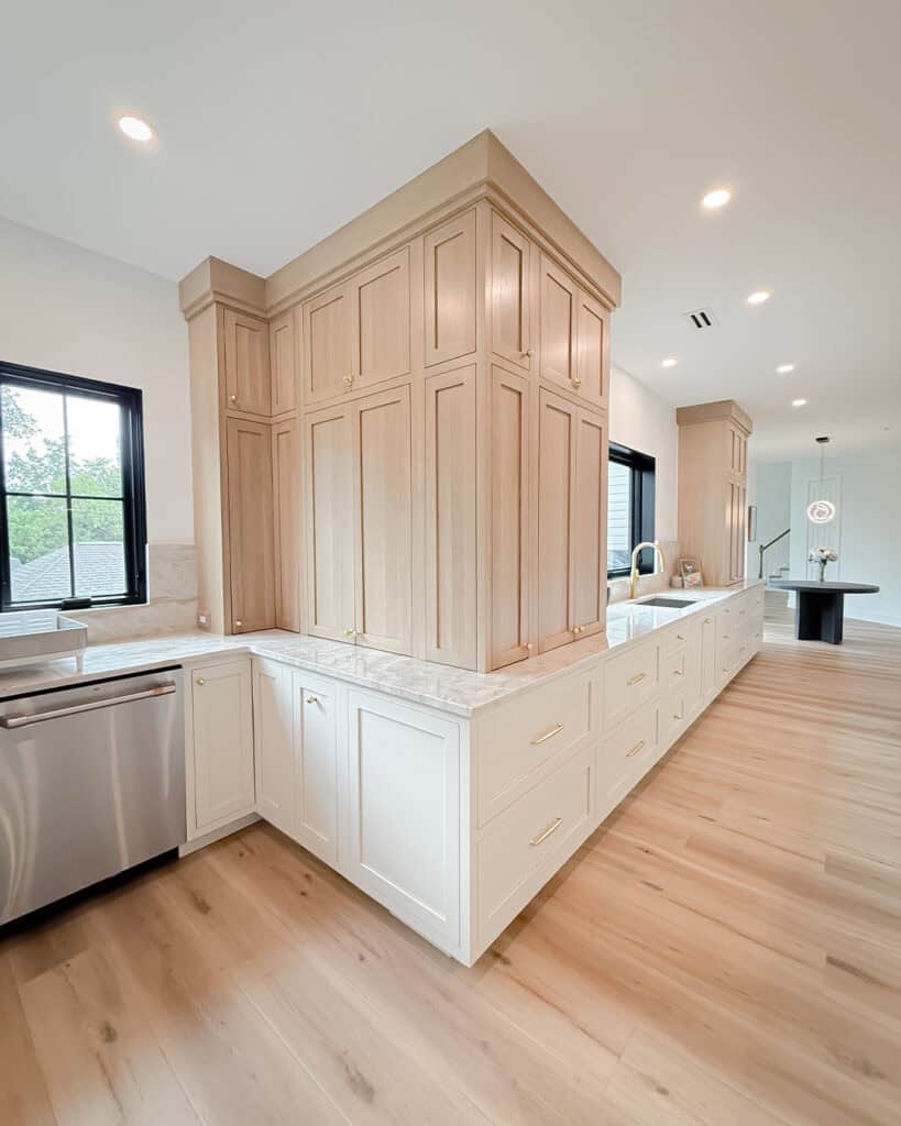 Modern kitchen with light wood cabinets, white marble countertops, built-in dishwasher, and light hardwood floors. Large windows provide natural light, creating a bright and spacious atmosphere.