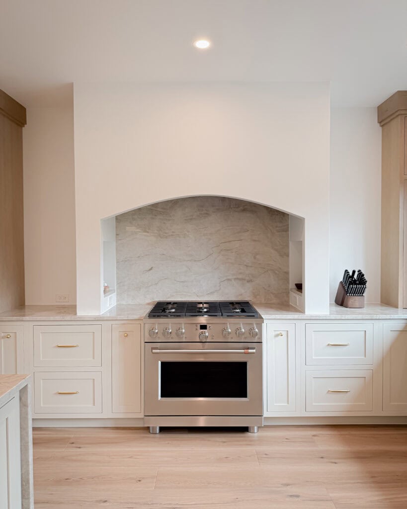 A modern kitchen features a stainless steel oven and gas stove set into white cabinetry, with a marble backsplash and light wood floors. A knife block sits on the countertop to the right.