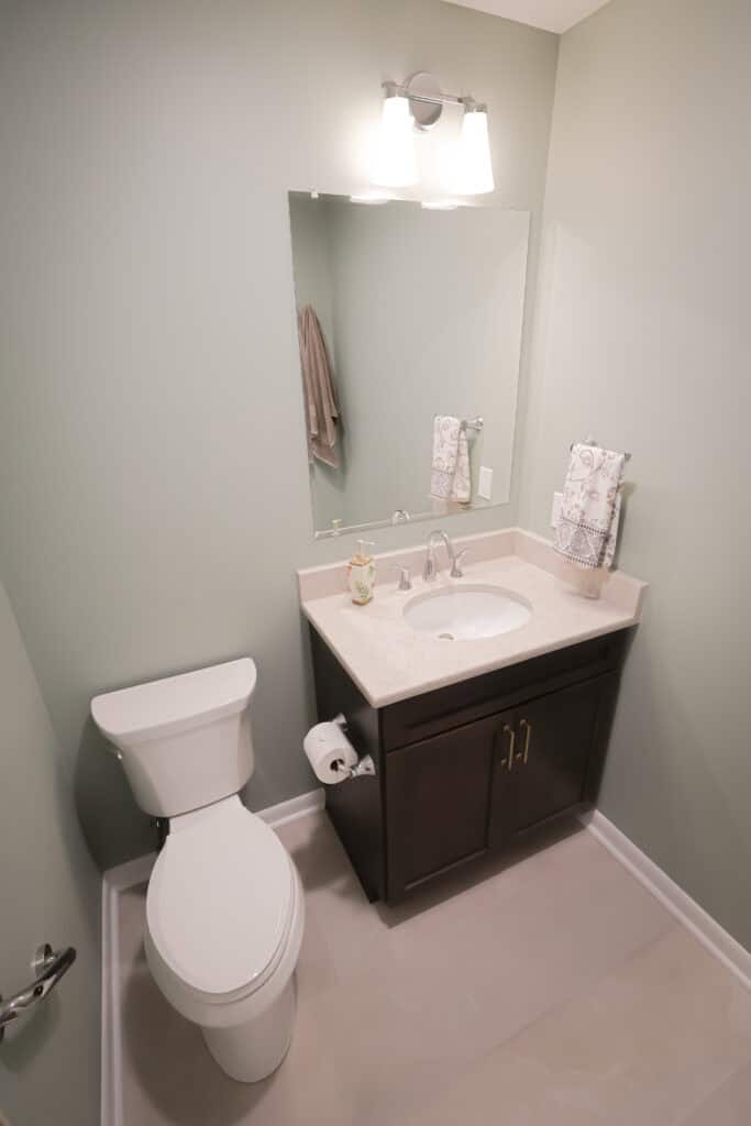 A small, modern bathroom with light green walls, a white toilet, a dark wood vanity with a white countertop, a rectangular mirror, and overhead lighting. A hand towel and soap dispenser sit on the counter.