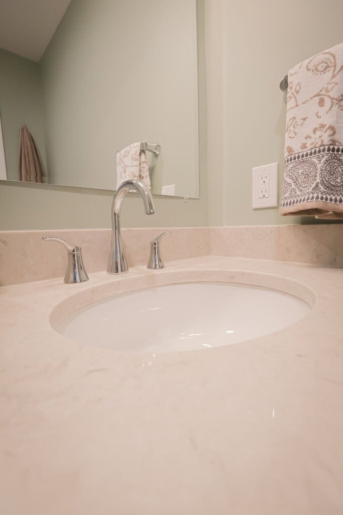 A modern bathroom sink with a polished chrome faucet, beige marble countertop, and a patterned towel hanging on a silver holder against a light green wall. A large mirror is visible in the background.