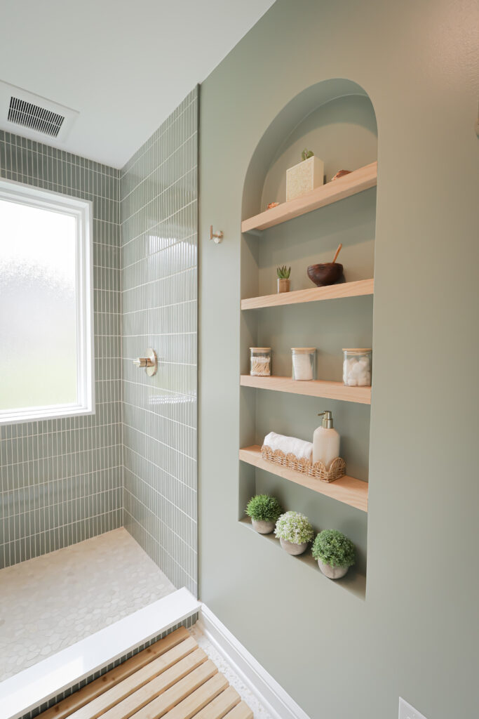 modern bathroom with green walls, a tiled shower, a window, and built in shelves holding jars, rolled towels, small plants, and decorative items. the space appears clean and minimalistic.