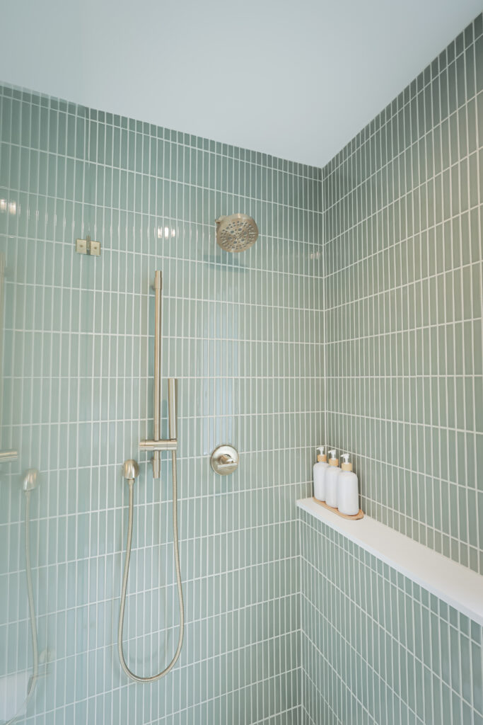 modern shower with vertical light green tiles, a rainfall showerhead, a handheld shower, and a white shelf holding four bottles. the design is clean and minimalistic.