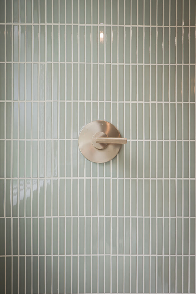 a silver shower handle on a wall with vertical, rectangular pale green tiles and white grout.