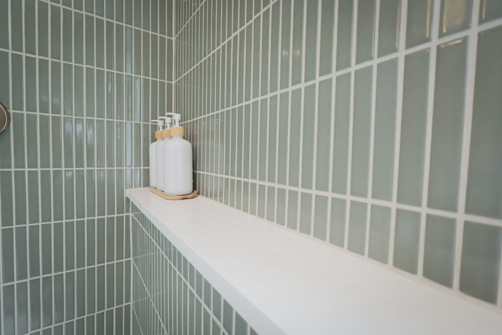 a white soap dispenser with a wooden top sits on a white shelf against a wall of green rectangular tiles in a bathroom.