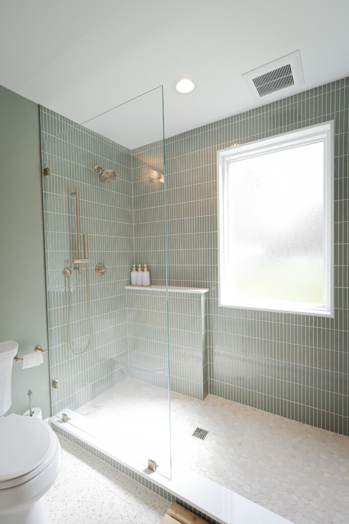 modern bathroom with a glass enclosed shower, vertical light green tiles, a built in shelf with bottles, and a frosted window. a white toilet is visible on the left.