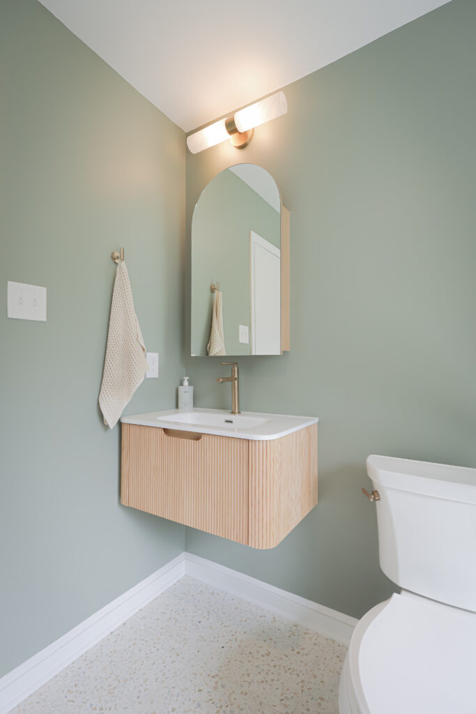 minimalist bathroom with sage green walls, a wall mounted wooden vanity, rectangular mirror with rounded top, modern light fixture, white toilet, textured hand towel, and terrazzo look flooring.