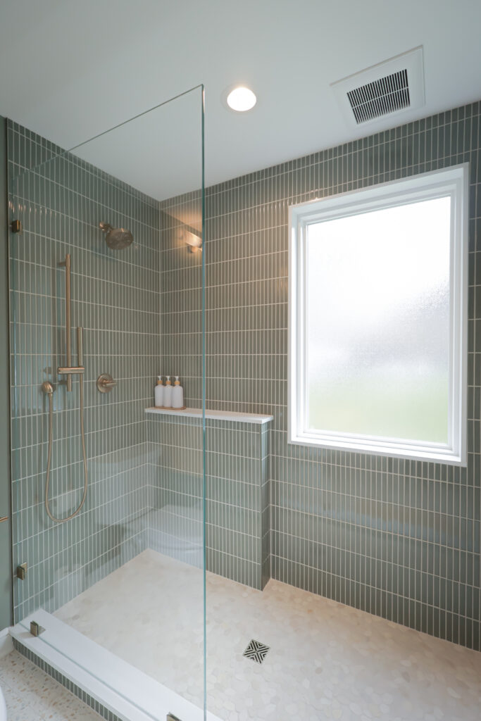 modern shower with green vertical tile, gold shower fixtures, a glass door, built in shelf holding bottles, and frosted window letting in light. beige hexagonal floor tiles complete the clean, minimalist look.