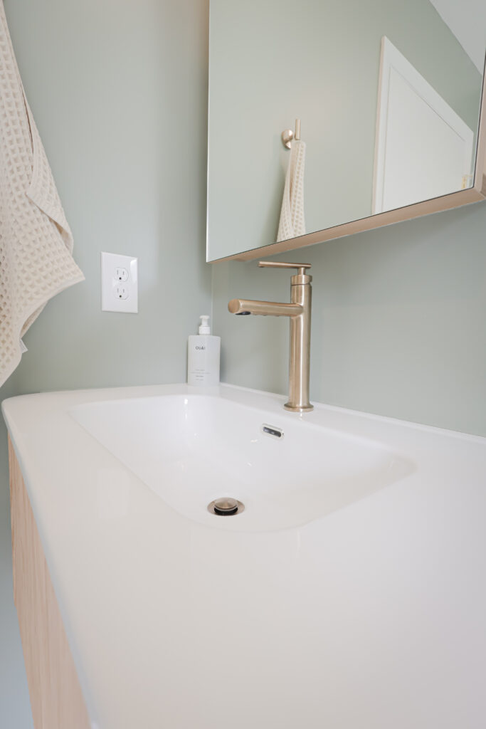 modern bathroom sink with a silver faucet, white countertop, soap dispenser, and a large mirror above. a textured towel hangs on the wall next to a white electrical outlet. the wall is painted a soft green.