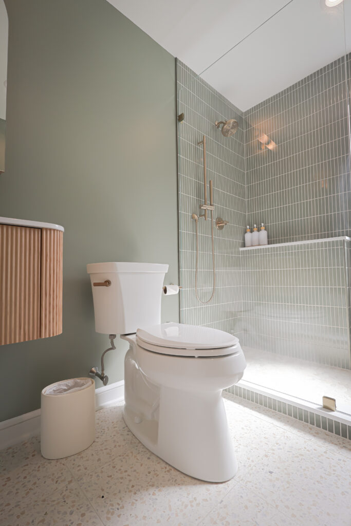 modern bathroom with a white toilet, a small trash can, green walls, a wooden vanity, and a glass enclosed shower with vertical green tiles and three bottles on a shelf inside.