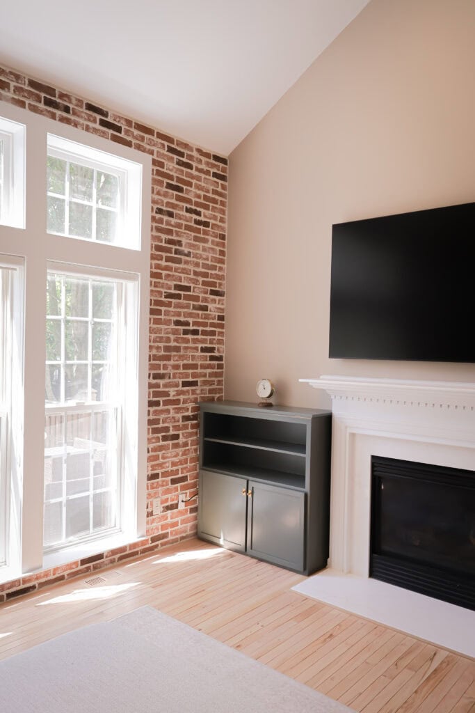a bright living room with large windows, an exposed brick wall, a black cabinet with a clock on top, a white fireplace, and a mounted flat screen tv above the mantel. sunlight streams in through the windows.